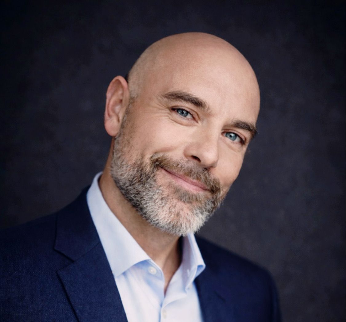 Oliver Charles Grann, stifter af Knowledge Nexus. Studieportræt i mørk jakke, smilende.